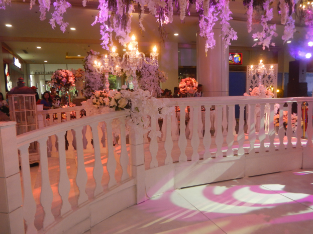 Weiße Reling mit Blumen und Kronleuchtern geschmückt, umgeben von Menschen, Säulen und Lichtern im Hintergrund und ein Bildschirm auf der rechten Seite.