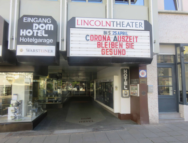 Außenansicht des Lincoln Theaters in Berlin, Deutschland, mit Glasfenstern und -türen und einer Tafel mit Text sowie einem belebten Stadtbild im Inneren.