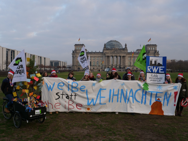 Gruppe von Menschen mit Mützen, die ein Banner vor dem Reichstaggebäude halten, mit einer Person auf einem Kinderwagen, grasbewachsenem Boden, Bäumen, Gebäuden und Fahnenmasten im Hintergrund unter einem bewölkten Himmel.