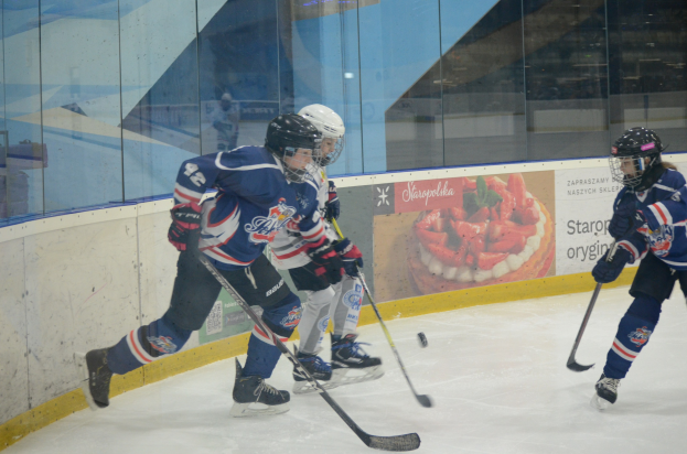 Gruppe junger Menschen, die Eis hockey auf einer Indoor-Eisfläche spielen, mit Helmen, Sportbekleidung und Hockey-Schlägern, mit einem Plakat im Hintergrund an der Glaswand.