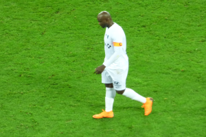 Ein Fussballspieler in einem weissen T-Shirt, weissen Shorts und orangen Schuhen läuft über ein saftiges grünes Feld.