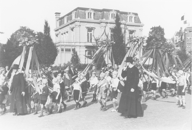 Schwarz-weißes Bild einer Gruppe von Menschen, die eine Straße entlanggehen und Fahnen vor einem Gebäude mit Bäumen und anderen Gebäuden im Hintergrund halten.
