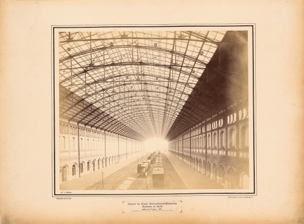 Schwarz-weißes Foto eines Bahnhofs mit einem Zug auf den Schienen, umgeben von Gebäuden mit Fenstern und einem Dach, das von Metallpfosten gestützt wird; Text ist unten im Bild sichtbar.