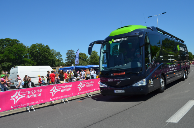 Ein schwarzer und grüner Bus fährt auf einer Straße neben einer Menge Menschen, einige tragen Mötzen, mit einem Banner auf der linken Seite und Bäumen unter einem klaren blauen Himmel im Hintergrund.