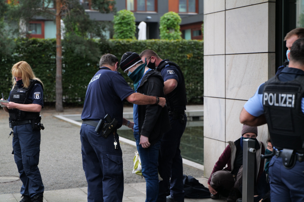 Eine Gruppe von Polizisten vor einem Gebäude stehend, mit einer Frau, die ein Buch und einen Stift hält, und mehreren Menschen in der Nähe sitzend und stehend, vor dem Hintergrund von Pflanzen, Bäumen und Gebäuden.