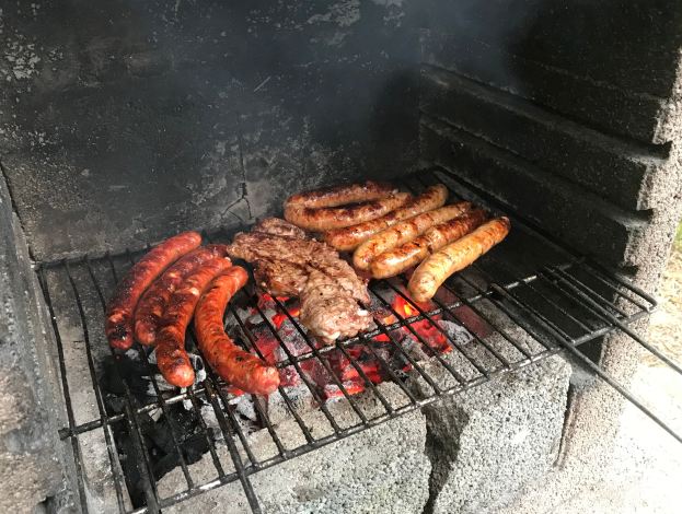 Ein Grill mit Würstchen und Fleisch darauf, umgeben von Kohle und Feuer, auf Gras im Freien.