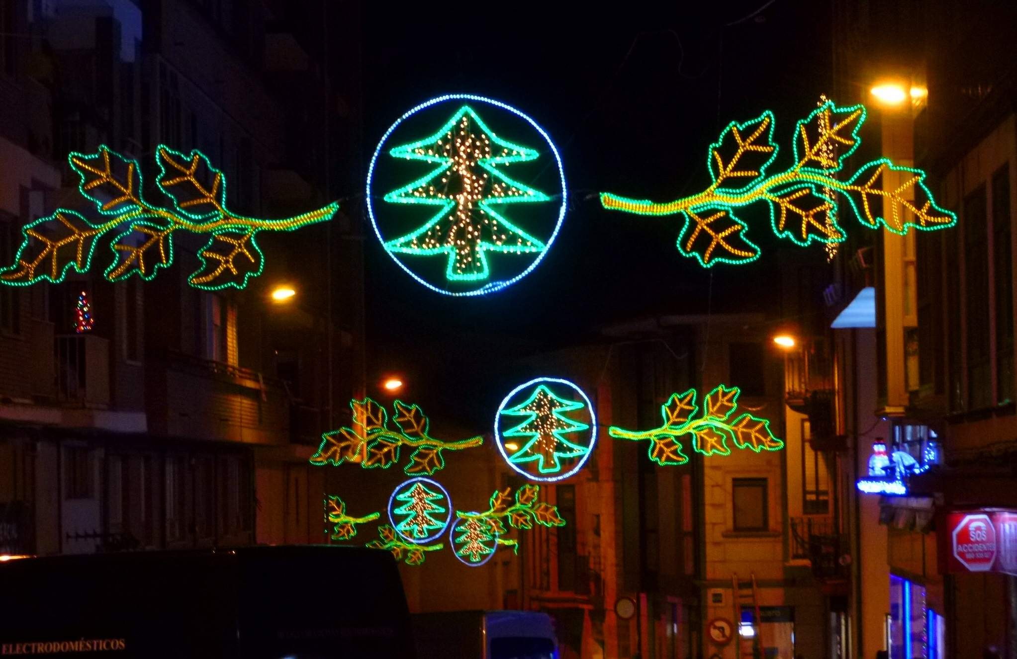 Eine nächtliche Stadtstraße, geschmückt mit festlichen Weihnachtslichtern mit Blättern und anderen Dekorationen, mit fahrenden Fahrzeugen und Gebäuden auf beiden Seiten.