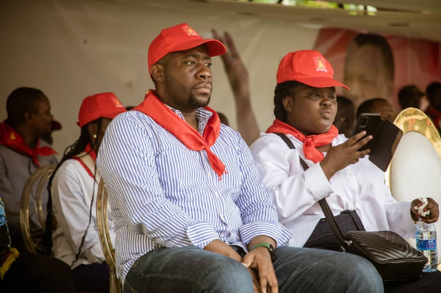 Eine Gruppe von Menschen in roten Mützen und Schals sitzt auf Stühlen unter einem Banner mit Lichtern darüber, einer hält ein Handy und ein Buch, ein anderer eine Tasche und eine Flasche, möglicherweise auf einer Veranstaltung der Demokratischen Partei.
