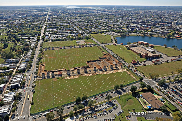 Luftaufnahme einer Stadt mit Gebäuden, Bäumen, Gras, Straßen mit Fahrzeugen, Wasser und Wolken, die einen großen zentralen Platz zeigt, der als Park geplant ist.