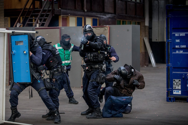 Eine Gruppe von Polizisten in Einsatzausrüstung steht vor einem Gebäude, einige halten Waffen, mit einer Person im Vordergrund und einem blauen Objekt mit Text auf der rechten Seite.