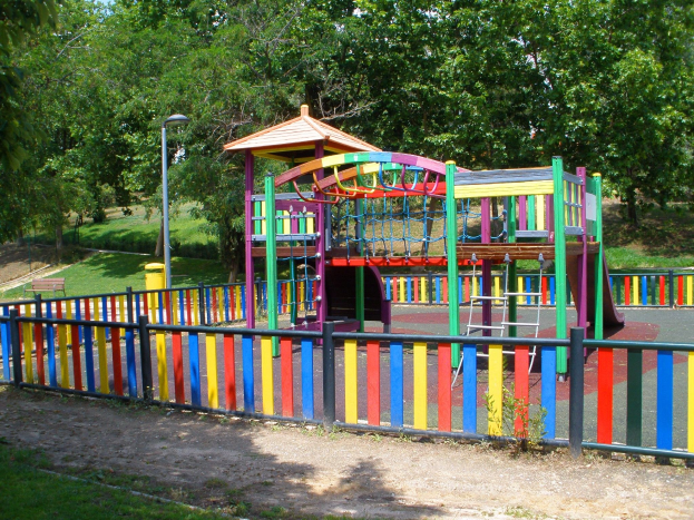Ein Spielplatz mit farbenfrohem Spielgerüt, umgeben von einem Zaun, Pfosten, Lichtern, Bänken, Gras, Pflanzen und Bäumen unter einem klaren blauen Himmel.