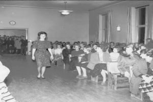 Eine Frau in einem schwarzen und weißen Kleid läuft über einen Laufsteg während einer Modenschau für Frauenwahlrecht, mit einer Menge, die auf Stühlen sitzt und zusieht, einer Uhr und Lampen an der Wand und einer beleuchteten Decke.