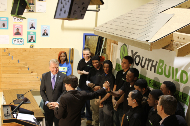 Gruppe von Menschen, die sich die Hände schütteln, um einen Podium bei einer öffentlichen Veranstaltung mit Mikrofonen und Papieren auf einem Tisch links, Plakate an der Wand und ein Objekt rechts.
