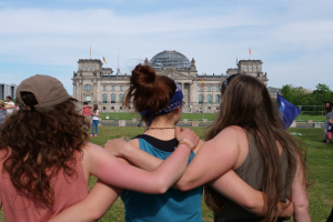 Drei Frauen in bunten Kleidern stehen vor dem Reichstag in Berlin, mit einer Menge, Flaggen und Bäumen im Hintergrund.