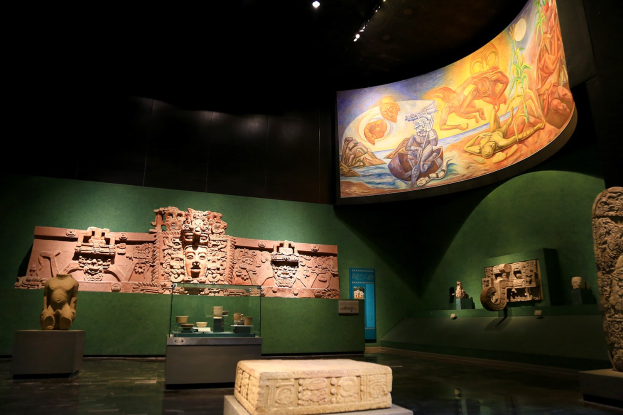 Ein Museumsraum mit verschiedenen Artefakten, darunter Skulpturen, Gegenstände auf Tischen, eine Texttafel, eine Wandmalerei und Deckenleuchten im Mexiko-Stadt-Museum der Anthropologie.