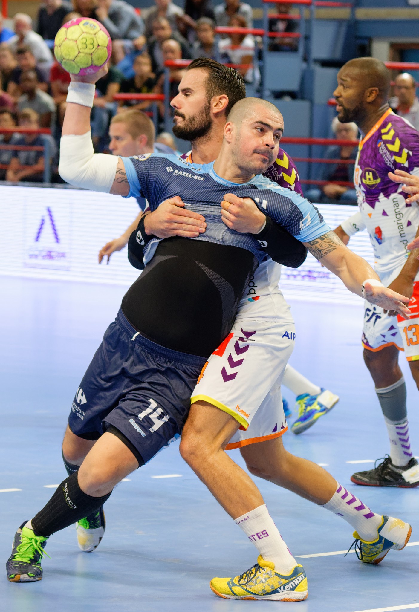 Mannergruppe spielt Hallenhandball mit einem Spieler, der den Ball hält, Zuschauern auf Tribüne und Geländer