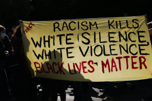Eine Gruppe maskierter Menschen steht auf einer Straße in der Nacht und hält ein Banner mit der Aufschrift 'Rassismus tötet Weiße Stille, Weiße Gewalt, Black Lives Matter'.