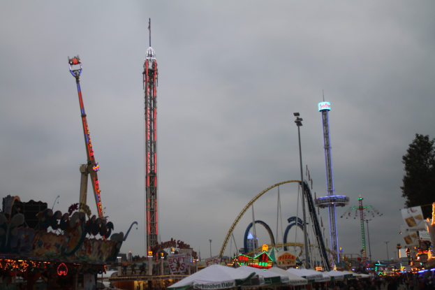 Eine Menschenmenge versammelt sich um einen hell erleuchteten Fasching bei Nacht, mit beleuchteten Fahrgeschäften, Pfählen und Bäumen unter einem bewölkten Himmel.