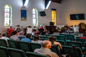 Eine Gruppe von Menschen sitzt auf Stühlen in einer Kirche, ein Mann steht vorne mit einem Mikrofon, umgeben von Musikinstrumenten, einem Tisch mit einer Blumenvase, einem Lautsprecher, einer Wand mit Text, Fenstern, einem Fernseher und Deckenleuchten.