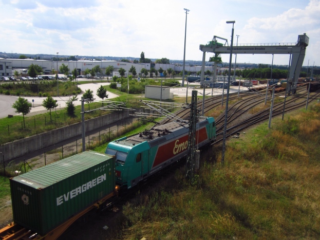 Güterzug fährt auf Schienen neben einem grünen Feld mit Strommasten, Kabeln, Bäumen, Gebäuden, Fahrzeugen auf der Straße und einer bewölkten Himmel.