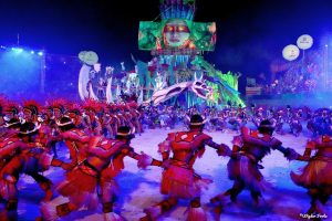 Eine Gruppe von Menschen in farbenfrohen Kostümen tanzt auf einer Bühne vor einem sitzenden Publikum, mit Lichtern, Stangen und Ballons, und ein Bildschirm zeigt eine Person im Hintergrund, während des Karnevals in Rio de Janeiro 2014.
