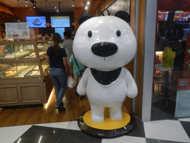 Eine Riesenpandastatue steht in einem Geschäft umgeben von Einkäufern, mit einer Auslage mit Lebensmitteln links und Fernsehgeräten an der Decke im Hintergrund.