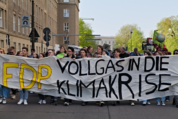 Eine Gruppe von Menschen marschiert auf einer Straße, die ein Banner trägt, auf dem "FDP Vollgas in die Klimakrise" steht, mit Gebäuden, Bäumen und Verkehrszeichen im Hintergrund unter einem klaren blauen Himmel.