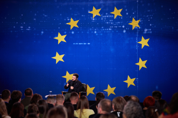 Ein Mann sitzt auf einem Stuhl vor einem Publikum, hält ein Mikrofon in der Hand, mit einem Glas und einer Flasche auf dem Tisch daneben und einem blauen Bildschirm mit gelben Sternen im Hintergrund.
