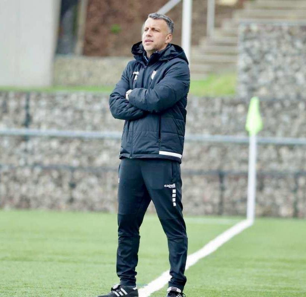 Ein Mann in schwarzer Jacke und Hose steht auf einem Fußballfeld mit verschränkten Armen und sieht konzentriert aus.