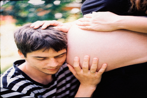 Ein Mann und eine Frau stehen Seite an Seite, der Mann berührt sanft den Bauch der Frau, mit einem unscharfen Hintergrund, der das Paar und ihr ungeborenes Kind hervorhebt.