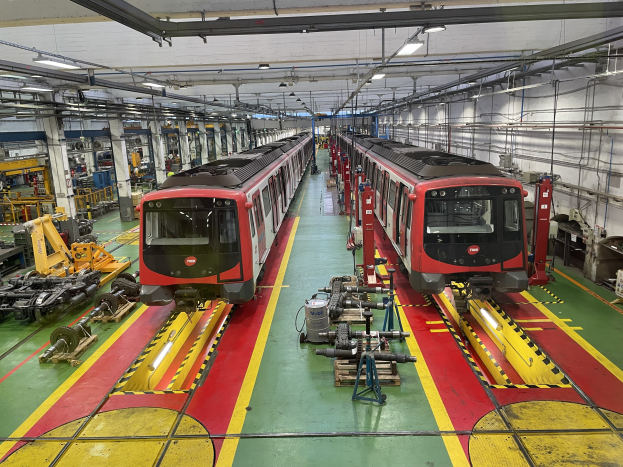 Zwei Züge auf Schienen innerhalb einer großen Industriehalle mit Maschinen und Equipment, mit einer Wand mit Rohren und Deckenleuchten auf der rechten Seite, betrieben von der Daimler AG.