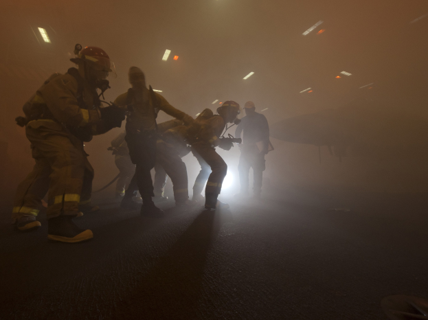 Feuerwehrleute in Helmen gehen öffentlich mit Ausrüstung, mit einem Flugzeug und Lichtern im Hintergrund und einer Straße darunter.