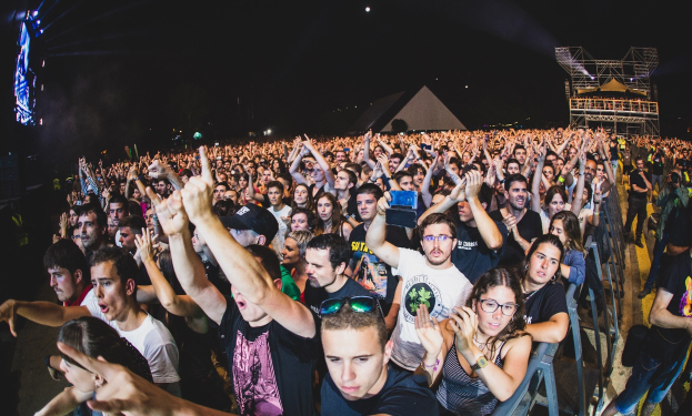 Menschenmenge auf einem Musikfestival mit erhobenen Händen, sitzend und stehend, hinter einem Metallzaun mit Gebäuden und Lichtern im Hintergrund.