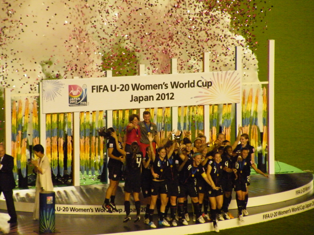 Eine Gruppe von Frauenfußballspielerinnen in schwarzen Trikots steht auf einer Bühne und feiert ihren Sieg, umgeben von einer Menge mit Kameras und einem dekorierten Hintergrund sowie einem grasbewachsenen Boden.