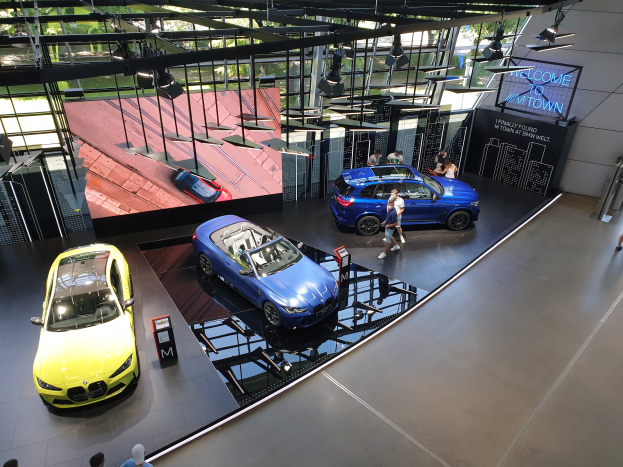 Eine Gruppe von Menschen steht im BMW Museum in München, mit Autos auf der linken Seite und einem Schild mit der Aufschrift "BMW Museum München" auf der rechten Seite, mit Blick auf Büsche, Geböude und den Himmel durch Glasfenster.