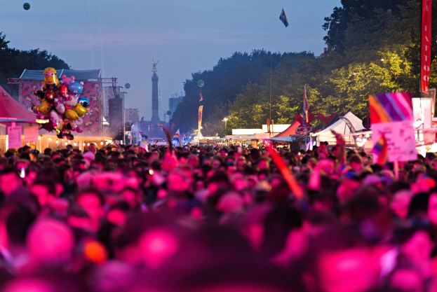 Eine große Menschenmenge auf einem nächtlichen Musikfestival, beleuchtet von bunten Lichtern, mit Zelten, Fahnen und Pfählen im Vordergrund, Bäume und ein Turm im Hintergrund unter einem klaren Himmel.
