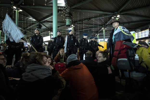 Eine Gruppe von Menschen steht vor einer größeren Menge auf einem Bahnhof, einige tragen Mützen und tragen Taschen, während andere Protestschilder und Banner halten unter Deckenleuchten und Säulen.