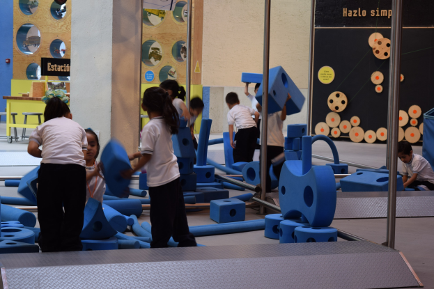 Kinder in einer Turnhalle, einige halten Gegenstände, mit einer Texttafel an der Wand und Stangen auf der rechten Seite.