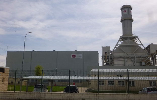 Großes Industriegebäude mit einem zentralen Turm, umgeben von einem Zaun, vor einer Straße mit Fahrzeugen, mit Pfählen, Lampen, Drähten, Bäumen und Gras im Vordergrund unter einem bewölkten Himmel.