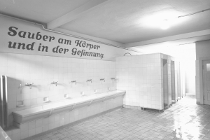 Schwarz-weiß-Foto einer öffentlichen Toilette mit Fliesenboden, Wandtext, mehreren Wasserhähnen, einer Tür und einer Deckenleuchte.