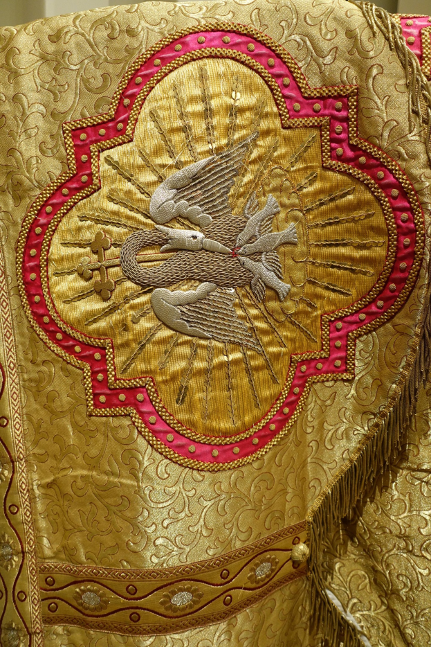 Nahaufnahme eines Priestergewandes mit aufwendiger Gold- und Rotstickerei und einem luxuriösen, königlich anmutenden Design vor einer Wand im Hintergrund.