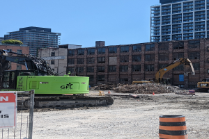 Eine Baustelle mit einem großen grünen Bagger in der Mitte, umgeben von einem Metallzaun und einem Schild an einem Pfahl, mit verschiedenen Maschinen, einem Eimer und einem Verkehrskegel im Vordergrund und Gebäuden, Bäumen und dem Himmel im Hintergrund.