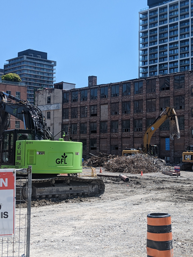 Eine Baustelle mit einem großen grünen Bagger in der Mitte, umgeben von einem Metallzaun und einem Schild an einem Pfahl, mit verschiedenen Maschinen, einem Eimer und einem Verkehrskegel im Vordergrund und Gebäuden, Bäumen und dem Himmel im Hintergrund.