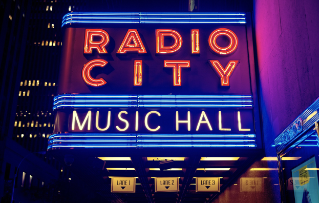 Außenansicht des Radio City Music Hall in New York City bei Nacht, hell erleuchtet und von hohen Gebäuden flankiert, mit einem "Radio City"-Schild und zusätzlichen Schildern auf der rechten Seite.