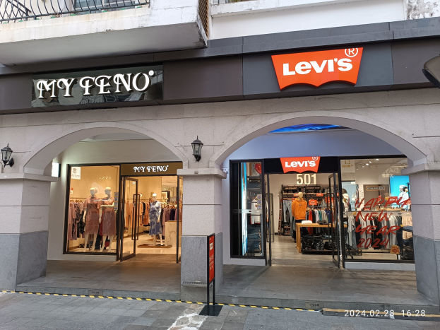 Ladenfront mit einem "Levi's"-Schild, Schaufensterpuppen in Kleidern, hängende Kleidung, Lampen, Texttafeln, Geländer und eine Straße unten.