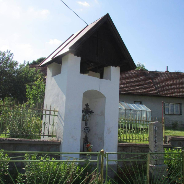Kleine weiße Kirche mit einem Kreuz darauf, umgeben von einem Metallzaun, Pflanzen und einem Weg, in der Nähe von Häusern unter einem bewölkten Himmel.