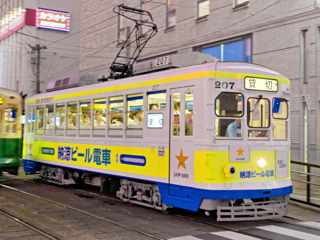 Eine gelbe und blaue Tram auf einer Stadtstraße bei Nacht mit Menschen drinnen, Gebäude mit Fenstern, Strommasten mit Drähten und Schilder mit Text im Hintergrund.