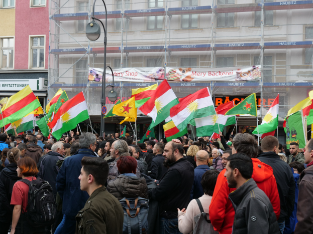 Große Gruppe von Menschen vor einem Gebäude mit einigen, die kurdische Flaggen halten, mit einem Laternenmast im Vordergrund und einer Banner mit Text im Hintergrund.