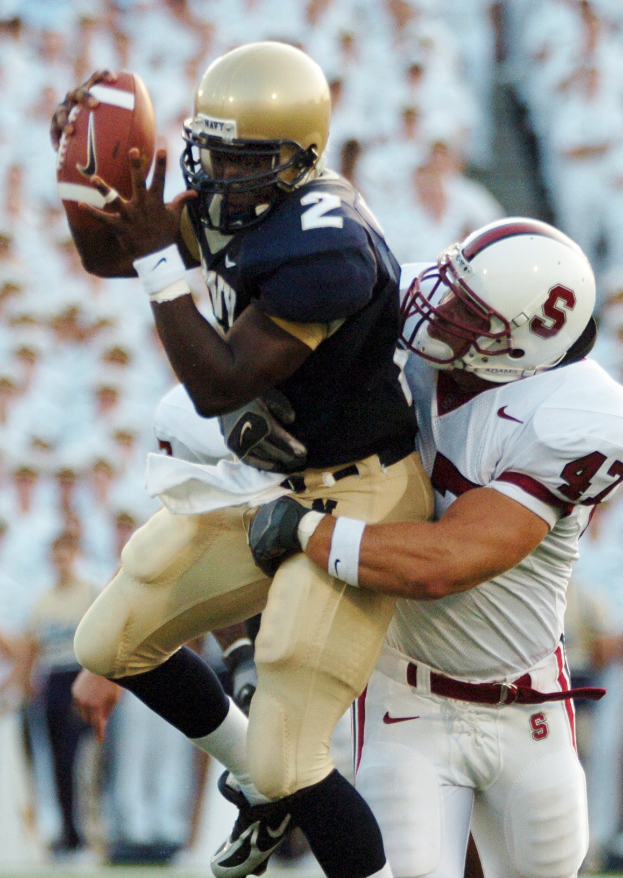 Zwei Fußballer in Helmen und Uniformen, einer fängt einen Ball in der Luft, während er von einem anderen getackelt wird, mit einem unscharfen Hintergrund.
