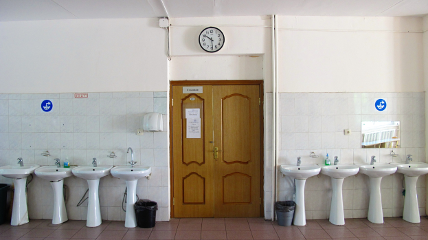 Ein Badezimmer mit einer Reihe von Waschbecken, Bodenmülltonnen, einer Tür, einer Wanduhr und einem Spiegel auf der rechten Seite.